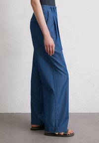 Wide-leg denim trousers in a medium blue shade, featuring front pleats, a button closure, and side pockets. Material appears soft and lightweight.