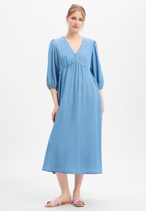Femme portant une robe midi bleu clair à col en V, manches trois-quarts bouffantes et sandales roses, debout sur un fond blanc uni.