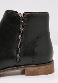 Bottine en cuir noir avec fermeture éclair latérale en métal et talon bas en bois marron, montrant des coutures détaillées sur une surface lisse.