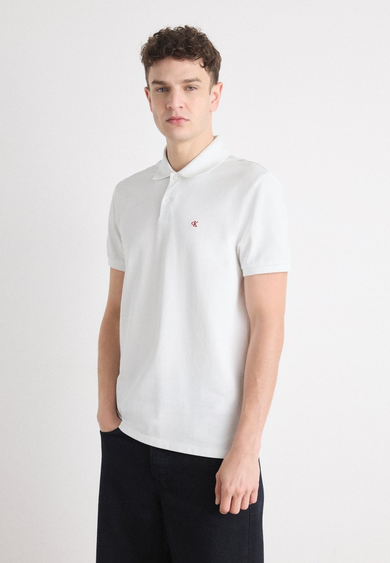 Hvid kortærmet polo shirt med spids krave, der har et lille rødt logo på venstre bryst og et blødt, textured stof.