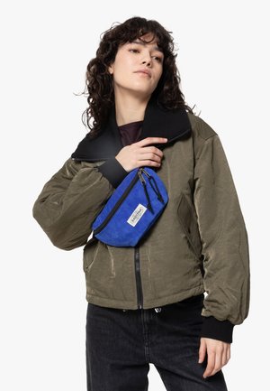 Olive green bomber jacket with a black collar, worn over a dark top, holding a blue corduroy pouch with a zipper and label.