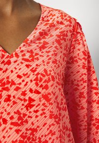Rot und rosa gemusterte Bluse mit V-Ausschnitt, glattem Stoff und einer Mischung aus einfarbigen und abstrakten Formen mit horizontalen Streifen.