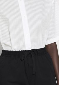 Camicia bianca a maniche corte con righe verticali, chiusa davanti con bottoni. Abbinata a pantaloni neri con vita elastica e cordoncino.