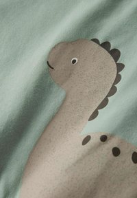 Tela de algodón en verde pálido con un diseño gráfico de un dinosaurio marrón con acentos negros. Textura suave, diseño simplista y formas redondeadas.