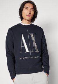 Felpa blu navy a maniche lunghe, colletto girocollo, con logo metallizzato argento "AX" e testo "ARMANI EXCHANGE" nella parte inferiore.