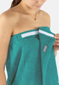 Türkiser Frottee-Handtuch mit weißem Klettverschluss und Etikett. Weiche, strukturierte Oberfläche, die sich um den Oberkörper legt.