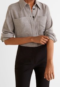 Camicia di lino grigia con colletto, due tasche sul petto e bottoni neri, indossata con pantaloni neri sartoriali, con le maniche arrotolate.
