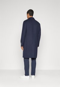 Marinblå ullrock med skräddarsydd design, lång silhuett, slagkrage och mönstrade knapprar; kombinerad med marinblå byxor och vita sneakers.