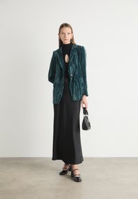 Teal velvet blazer with vertical stripes, black turtleneck featuring a keyhole, long black skirt, and black handbag; model wears black heels.