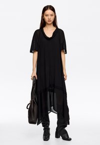 Musta epäsymmetrinen tunikamekko lyhyillä hihoilla, pehmeää kangasta, leveä kaula-aukko ja läpikuultava alahelma. Mallilla on ruskea laukku kädessään ja päällään mustat housut.