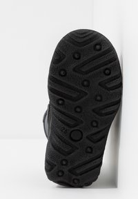 Semelle noire robuste d’une chaussure avec des crampons circulaires de traction et un motif de bande de roulement en zigzag texturé, sur fond blanc.