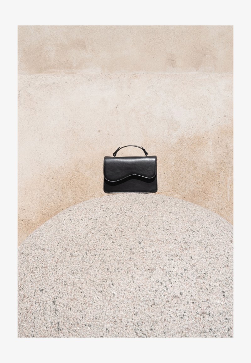 Sac à main en cuir noir avec rabat incurvé et poignée supérieure posé sur une grande sphère en pierre claire texturée, devant un mur beige.
