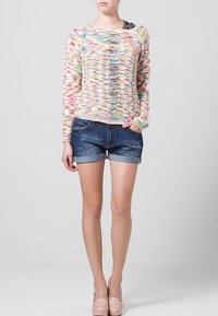 Suéter de punto colorido con cuello redondo y detalle de abertura en la parte frontal, combinado con pantalones cortos de mezclilla oscuros y zapatos de tacón nude.