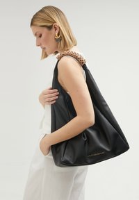 Sac à main en cuir noir avec un design souple et décontracté. Bandoulière tissée en tan et accent en logo doré. Texture lisse et intérieur spacieux.
