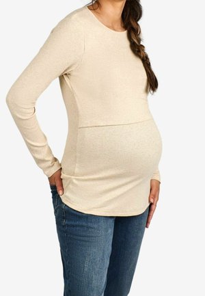 Zwangere vrouw in een beige top met lange mouwen en blauwe jeans, staand met één hand op haar heup en de andere rustend op haar buik.