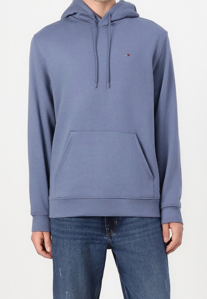 Homme portant un sweat à capuche bleu avec poche avant et cordons, associé à un jean bleu sur fond blanc.