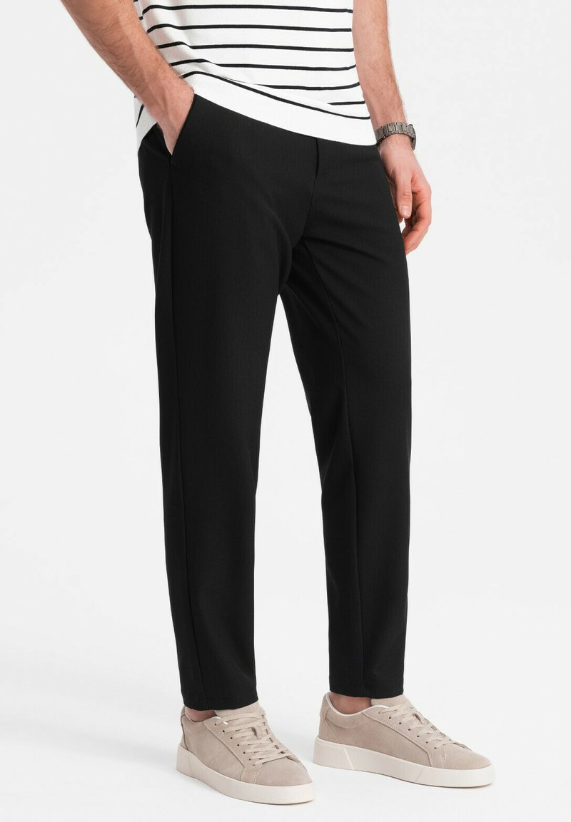 Zwarte, op maat gemaakte broek met een gladde textuur, voorzien van zijzakken en een rechte pasvorm. Gecombineerd met beige sneakers voor contrast.