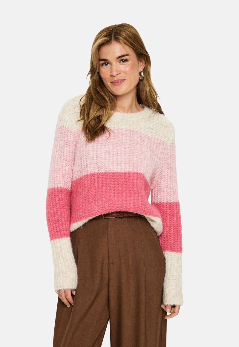 Kvinna med ljust brunt hår som bär en randig rosa och krämfärgad tröja instoppad i högmidjade bruna byxor med örhängen.