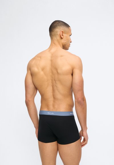 Μυώδης άνδρας στέκεται με την πλάτη προς την κάμερα, φοράει μαύρα αντρικά εσώρουχα τύπου μποξεράκια με γκρι ζώνη μέσης σε απλό λευκό φόντο.