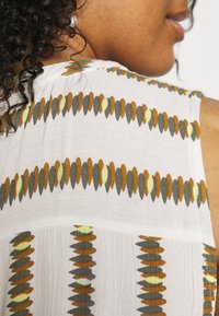 Top senza maniche in tessuto bianco leggero, caratterizzato da righe verticali di motivi ovali verdi, gialli e marroni sulla parte posteriore.