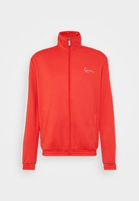 Chaqueta roja con cremallera hecha de una tela suave. Presenta un cuello alto, dos bolsillos laterales y un logo blanco en el lado izquierdo del pecho.