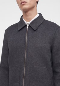 Homme portant un manteau en laine gris foncé zippé avec un large col par-dessus une chemise blanche boutonnée.