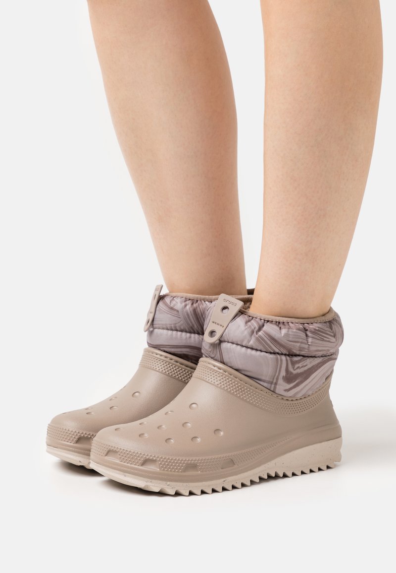 Botas impermeáveis bejas com uma sola texturizada, apresentando um cabedal de tecido com padrão e um punho acolchoado, projetadas para calor e conforto.