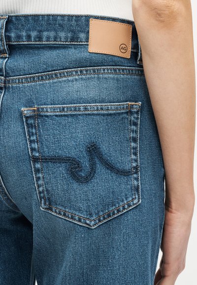 Πίσω μέρος μπλε τζιν με κεντημένο σχέδιο κύματος στην τσέπη και ταμπέλα από ταμπά δέρμα με το λογότυπο «AG», φορεμένα με λευκό ριμπ τοπ.