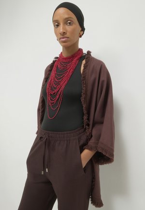 Brown fringed shawl over a black top, paired with brown pants. Multiple red beaded necklaces add texture and color, enhancing the outfit.
