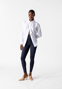 Biała koszula z kołnierzykiem założona na dopasowane ciemnoniebieskie legginsy, w połączeniu z beżowymi szpilkami, na jednolitym białym tle.