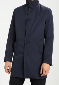 Cappotto blu navy con colletto alto, cinque bottoni frontali, due tasche laterali e tessuto testurizzato. Taglio aderente e maniche lunghe.