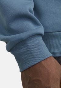 Détail d'un sweatshirt en coton bleu montrant un poignet côtelé ; texture lisse avec un design ajusté. Une main avec une peau texturée est visible.