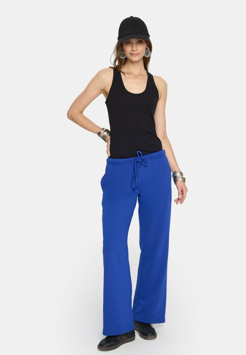 Kobieta stoi w czarnym topie bez rękawów, czapce w kolorze czarnym, srebrnych bransoletkach i kolczykach, jasnoniebieskich dresach i czarnych butach na białym tle.