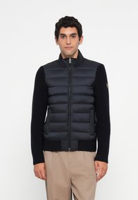 Homme portant un gilet matelassé noir avec des manches tricotées et un pantalon beige, se tenant devant un fond blanc uni.