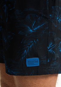Czarne szorty kąpielowe z wyrazistym niebieskim kwiatowym wzorem, z niebieską metką z napisem "Petrol Ind" przyszytą z boku. Miękki, teksturowany materiał.
