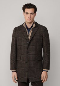 Abrigo de lana marrón a cuadros con solapas con muesca, dos botones y bolsillos laterales. Presenta un collar beige en capas y una tela texturizada.
