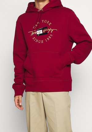 Man draagt een rode Tommy Hilfiger hoodie met een buidelzak en beige broek, staand met één hand in de zak.