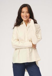 Cremefarvet skjorte med knapper og lange ærmer, afslappet pasform og buet kant. Har en lomme på brystet og blødt bomuldsstof.