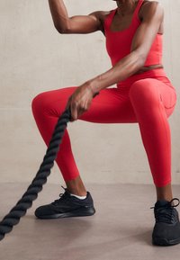 Czerwony stanik sportowy i legginsy w pozycji przysiadu, trzymające czarną linę treningową. Czarne buty sportowe dopełniają strój treningowy na neutralnej podłodze.