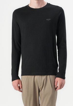 Homme portant un t-shirt noir à manches longues et col rond avec un petit logo "JOOP! JEANS" sur la poitrine, associé à un pantalon beige, debout sur fond blanc.