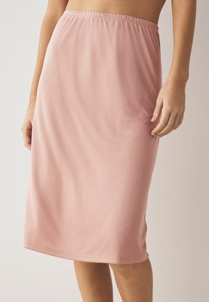 Mujer que lleva una falda rosa claro hasta la rodilla con textura lisa, de pie frente a un fondo claro y liso.
