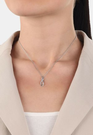Collar de plata con un colgante entrelazado, adornado con acentos similares a diamantes, colgando de una fina cadena. Modelo luciendo un blazer beige.