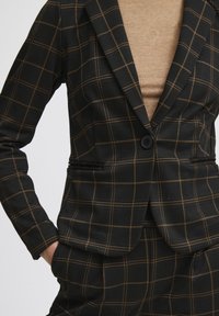 Blazer à carreaux noirs avec des lignes marron, design ajusté, fermeture à un bouton et deux poches avant ; porté sur un haut marron clair.