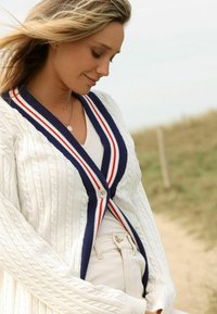 Kabelgebreide witte cardigan met een marineblauwe en rode gestreepte kraag, voorzien van een knoopsluiting en lange mouwen. Effen textuur en een ontspannen pasvorm.