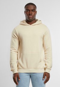 Felpa beige realizzata in tessuto morbido, dotata di tasca sul davanti, cappuccio con lacci e polsini a costine. Indossata con jeans azzurri chiari.