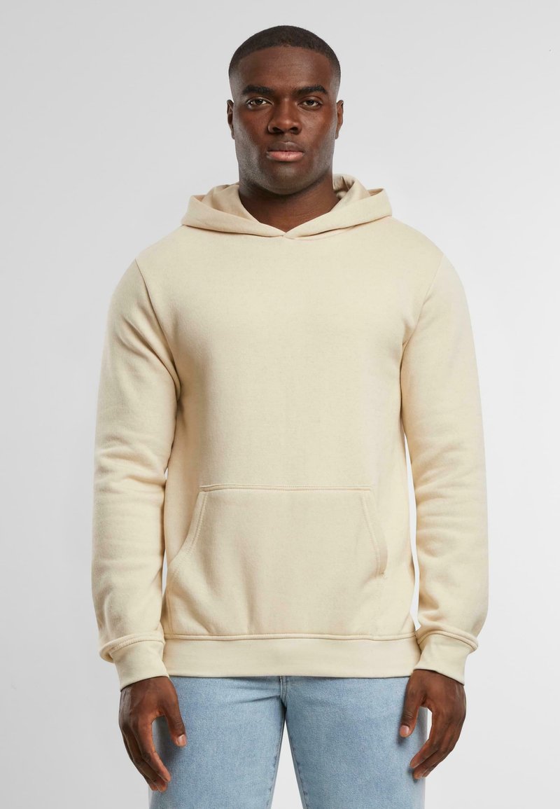 Felpa beige realizzata in tessuto morbido, dotata di tasca sul davanti, cappuccio con lacci e polsini a costine. Indossata con jeans azzurri chiari.