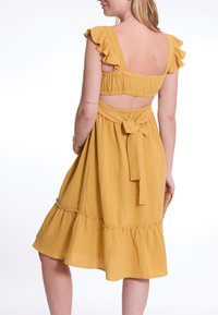 Senfgelbe, knielange Kleid mit einem rüschigen Ausschnitt, offener Rücken, gebundenem Taillenband und weit auslaufendem Rock mit gestuftem Saum. Weicher, strukturierter Stoff.