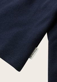 Primer plano de una tela azul marino que muestra el dobladillo de la manga con doble costura y la etiqueta de la marca Tom Tailor cosida en la costura lateral.