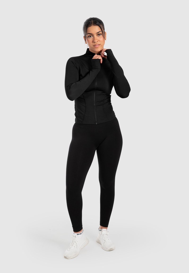 Schwarzes langärmliges Zip-Up-Oberteil und Leggings aus dehnbarem Material, kombiniert mit weißen Sneakern. Glatte Textur mit tailliertem Design.