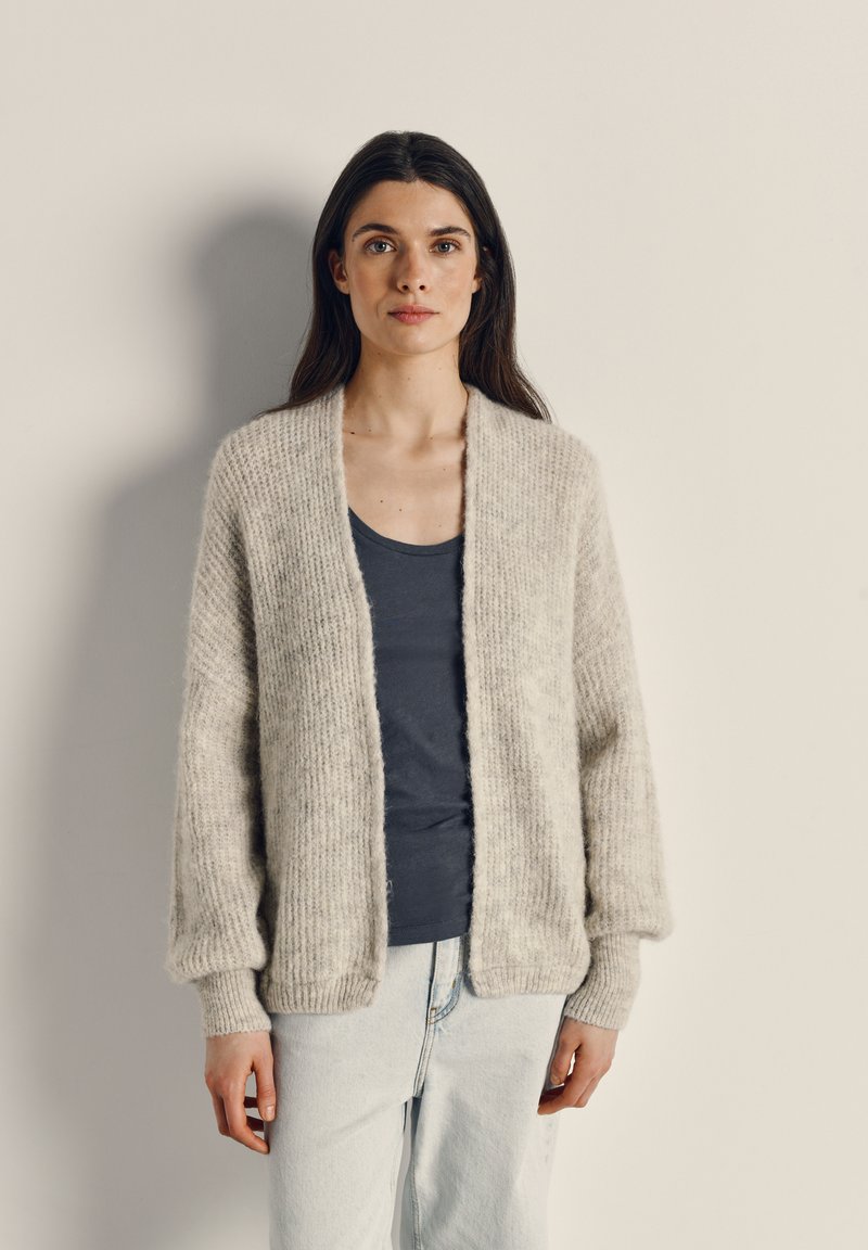 Hellgraue, grob gestrickte Strickjacke mit geripptem Muster, offener Front und umgeschlagenen Ärmeln, getragen über einem marineblauen Tanktop und hellen Jeans.